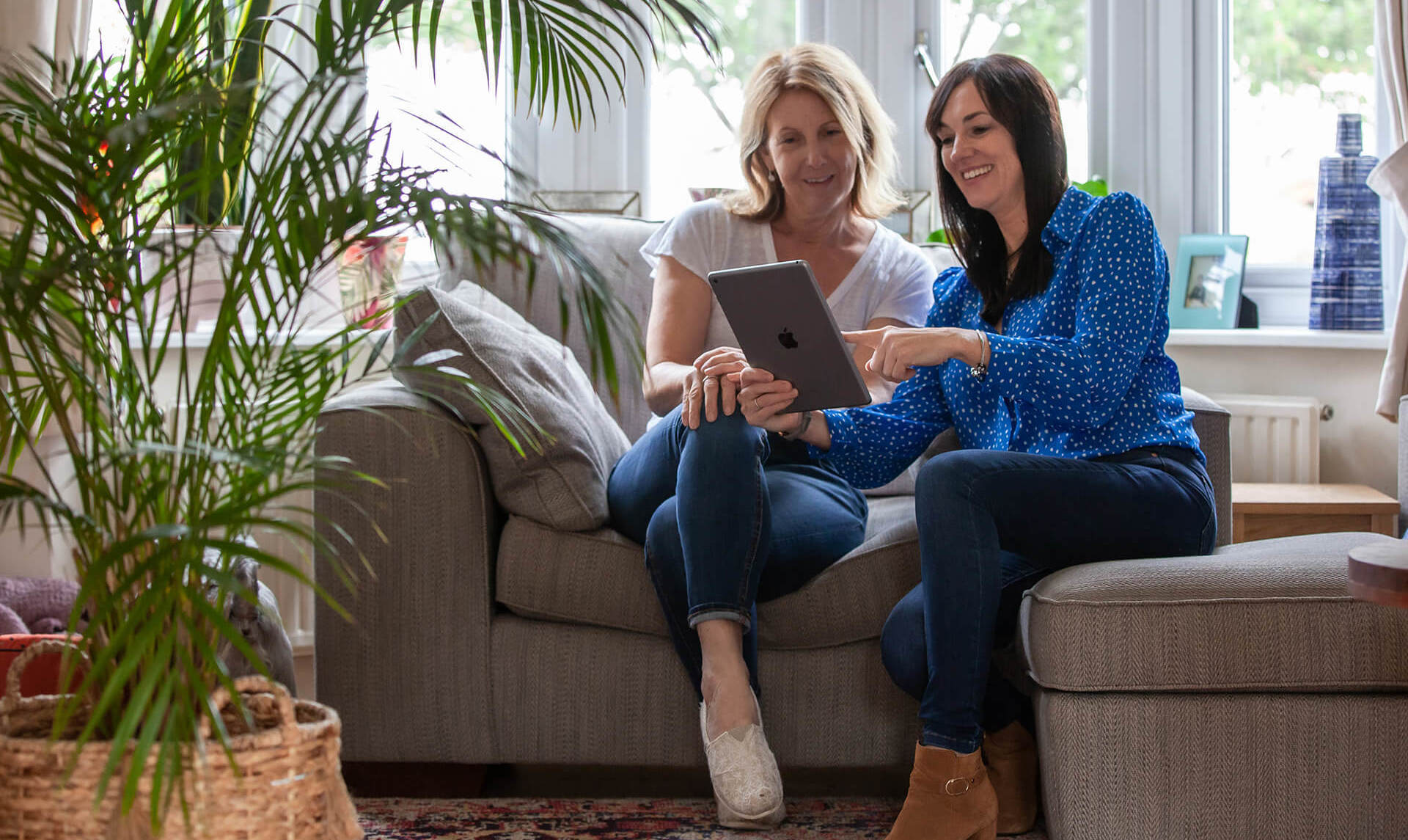 Two smiling women sat on a sofa looking at a screen on a tablet